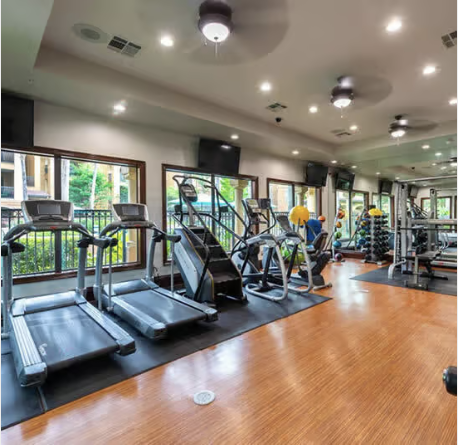 Francis Property Management Modern gym interior with treadmills, elliptical machines, and weights at Estancia San Miguel, featuring large windows and a wooden floor.