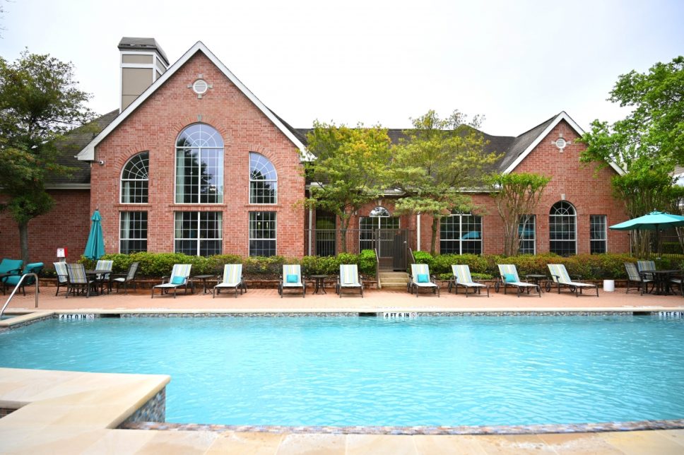 Francis Property Management The Bellagio Apartments in Houston, Texas, feature a swimming pool surrounded by lounge chairs, set in front of a large brick building with tall arched windows and trees around it.