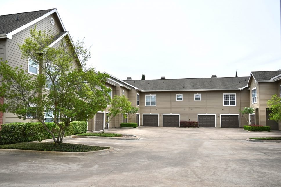 Francis Property Management Two-story residential buildings at the Bellagio Apartments in Houston, Texas feature garages on the first floor. The courtyard boasts parking spaces and is landscaped with trees and shrubs. The sky is overcast.